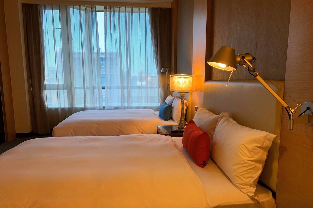 Ein Hotelzimmer mit zwei Betten, einem Nachttisch mit Lampe und Blick auf die Stadt durch transparente Vorhänge.