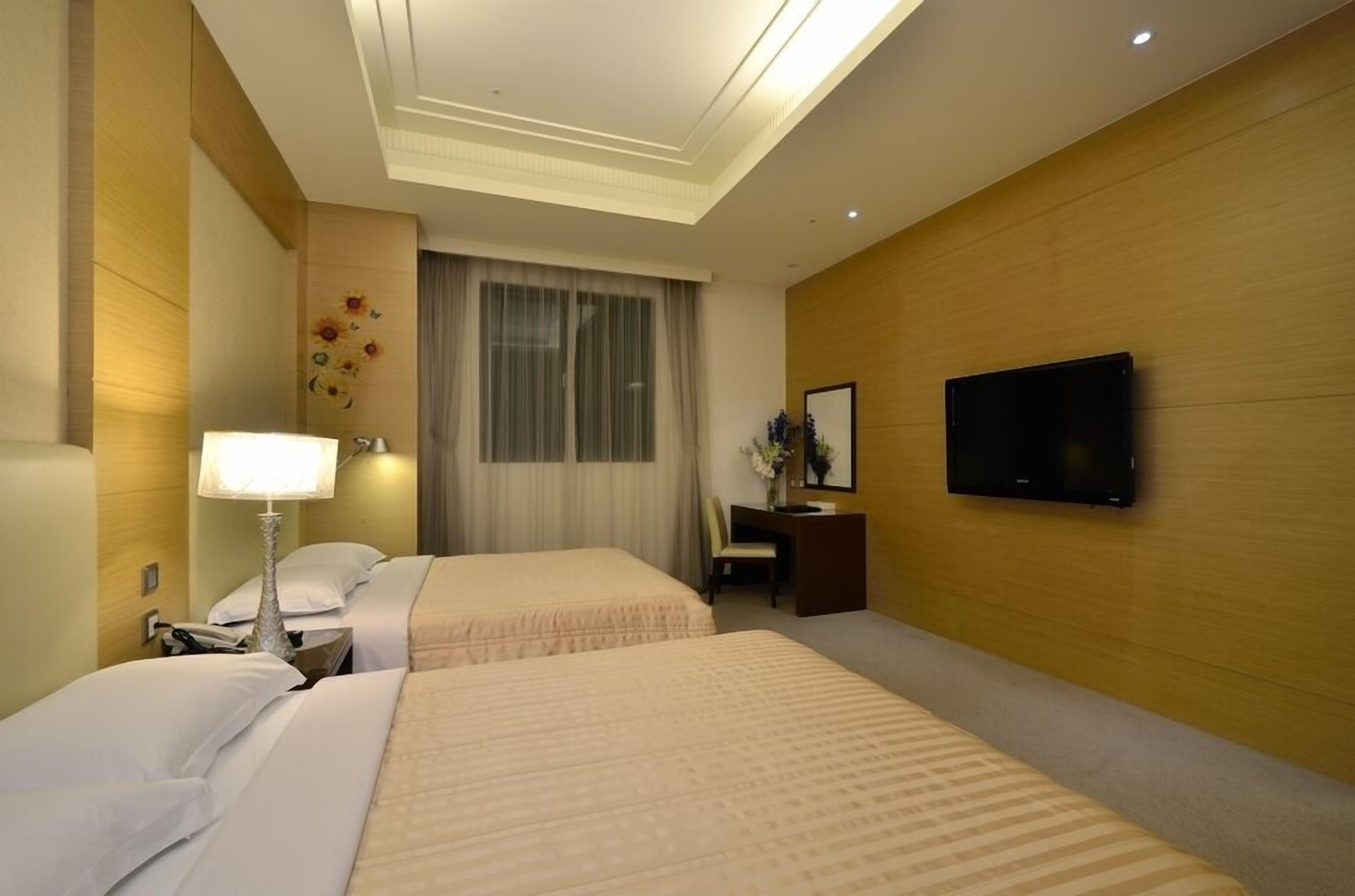 A hotel room with two beds, a desk with a chair, a flat-screen TV, and a window with curtains.