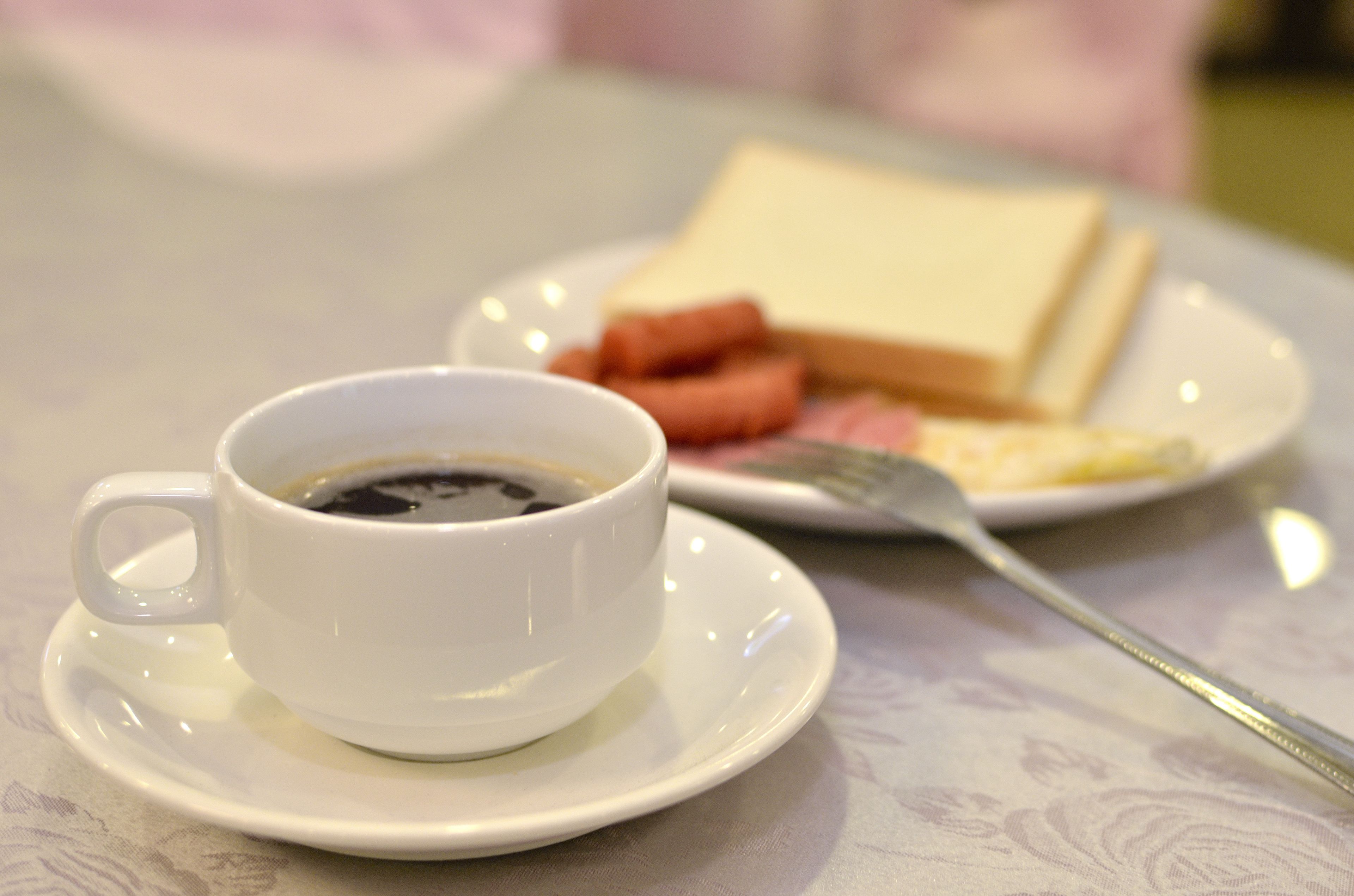 Een witte koffiekop op een schoteltje, een bord met toast en een vork, en een bord met worst en een stuk vlees.