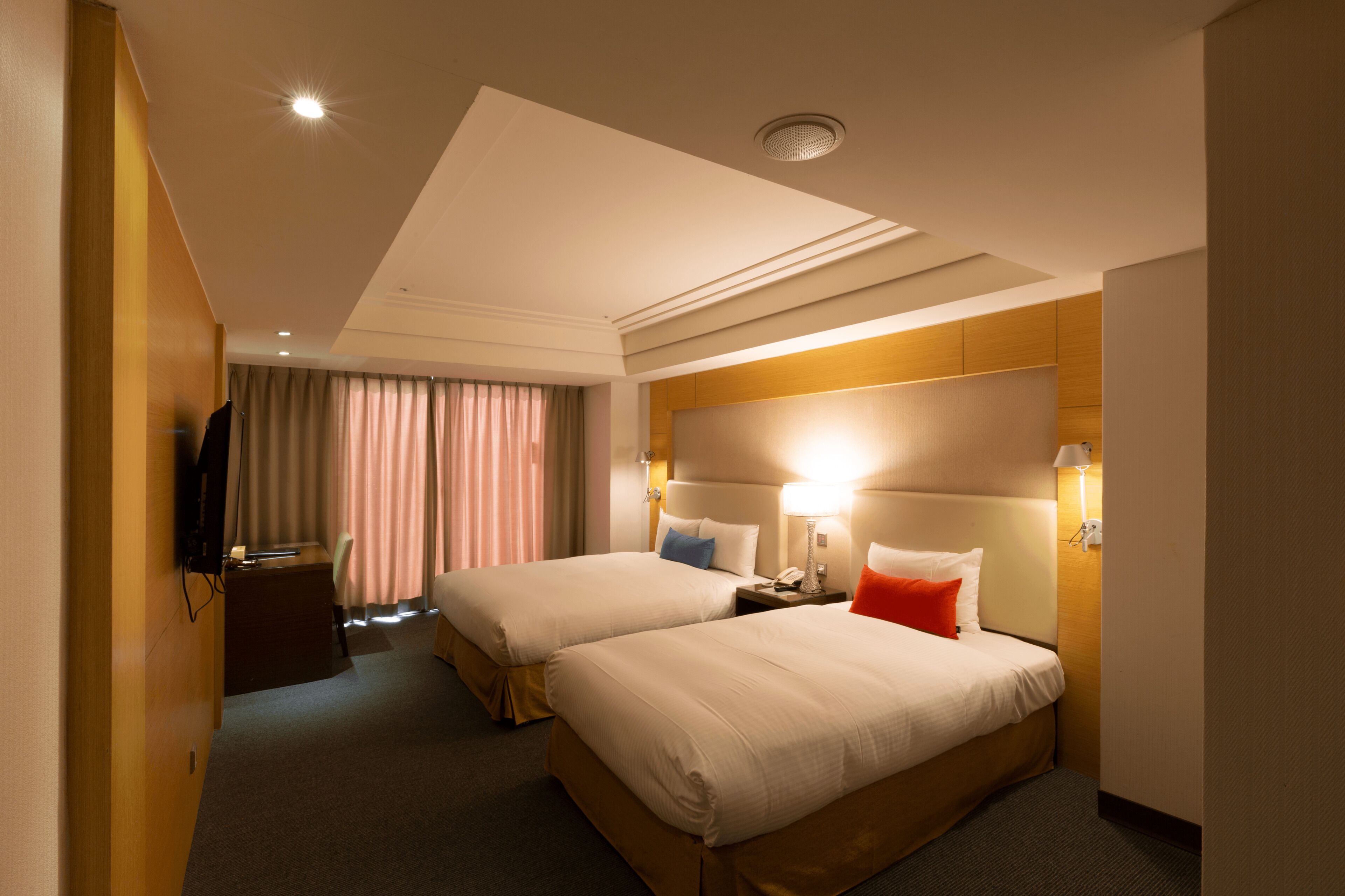 A hotel room with two beds, a desk, a television, and a window with curtains.