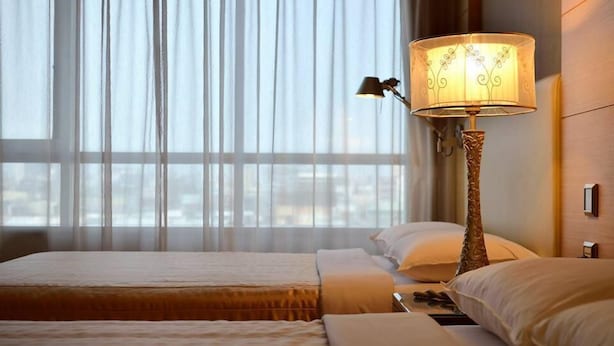 A hotel room with two beds, a lamp, and a view of the city through sheer curtains.