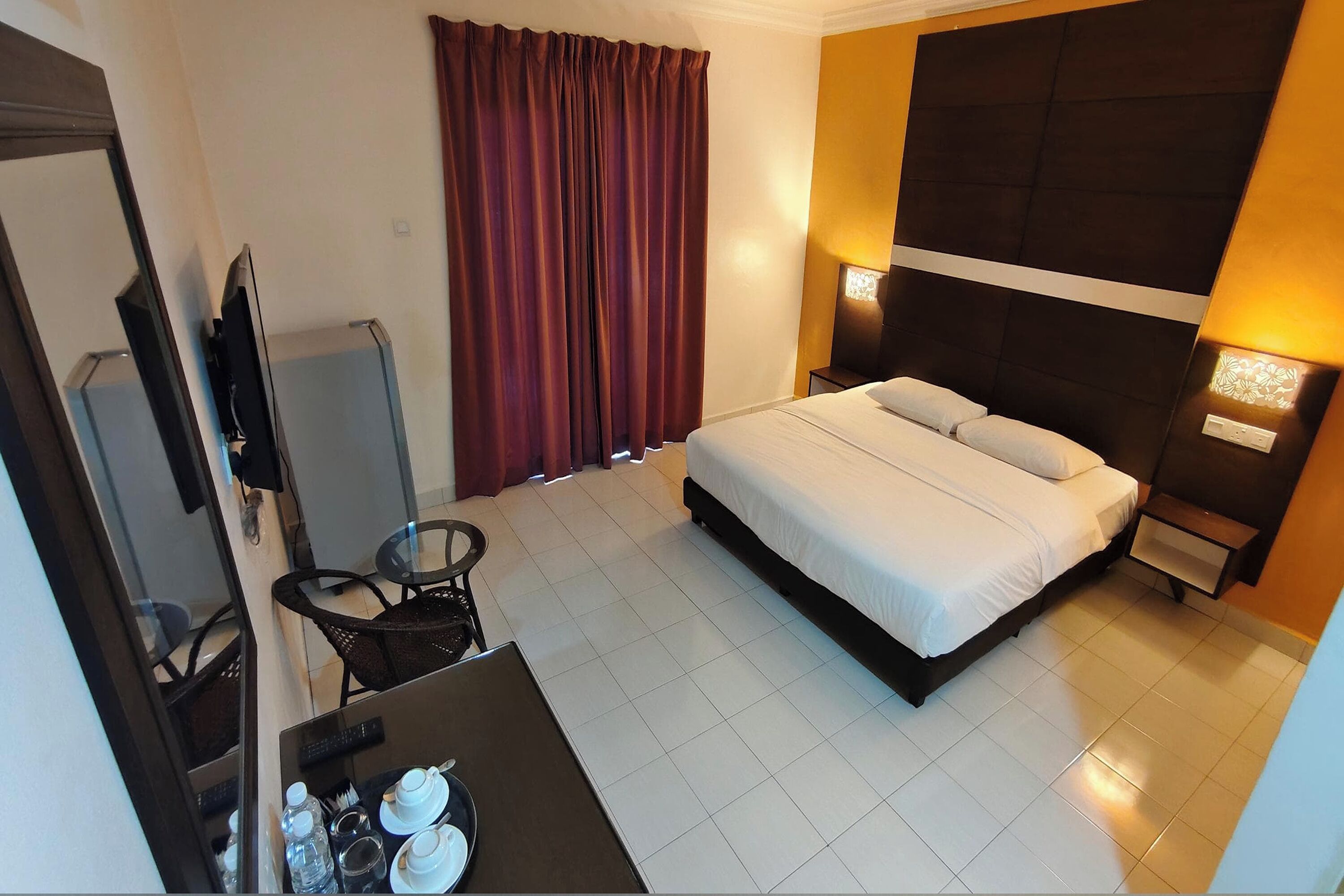 A hotel room with a bed, a TV, a desk with a chair, a table with cups and a bottle, and a window with curtains.