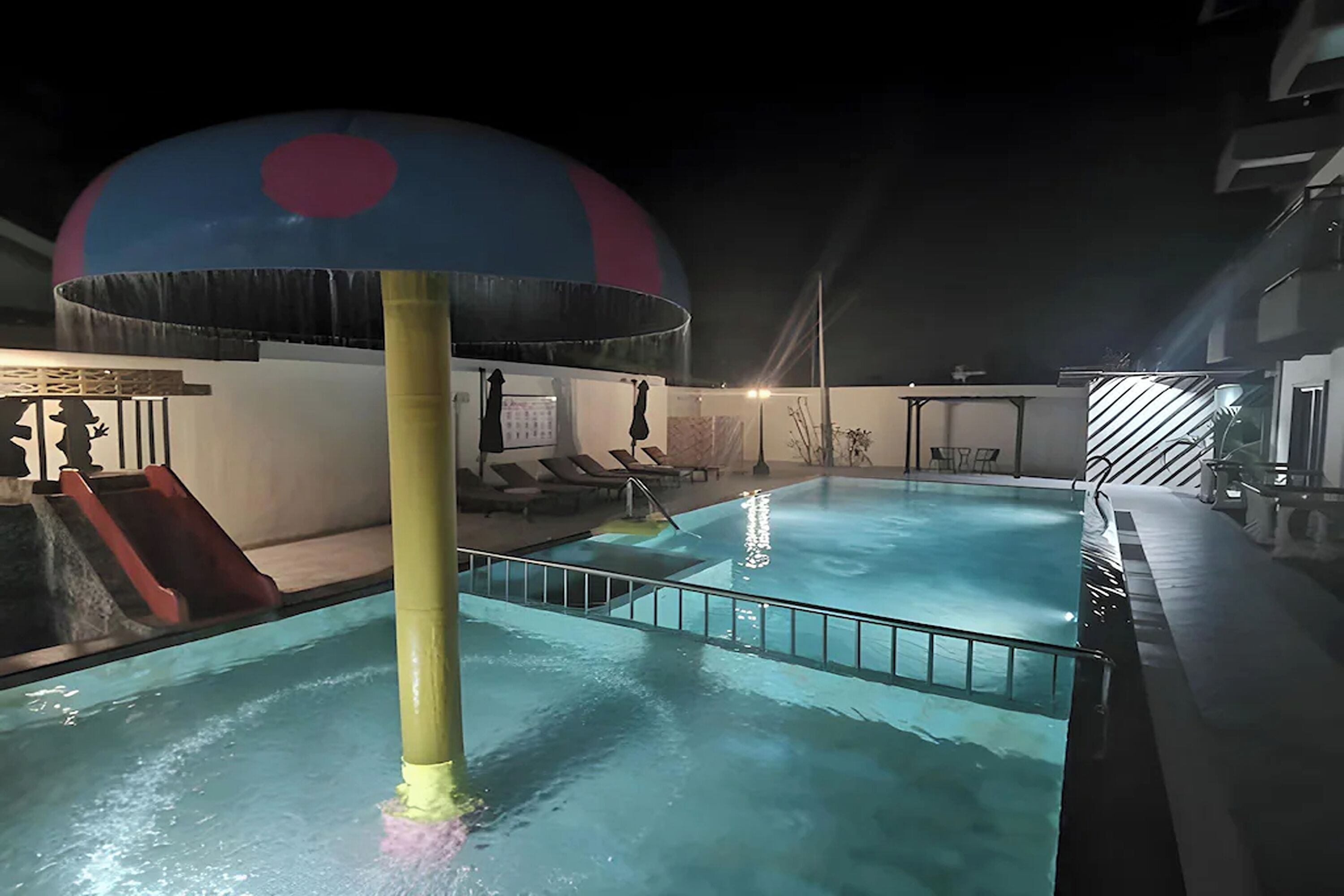 A swimming pool area with a slide, a large water play structure, and lounge chairs.