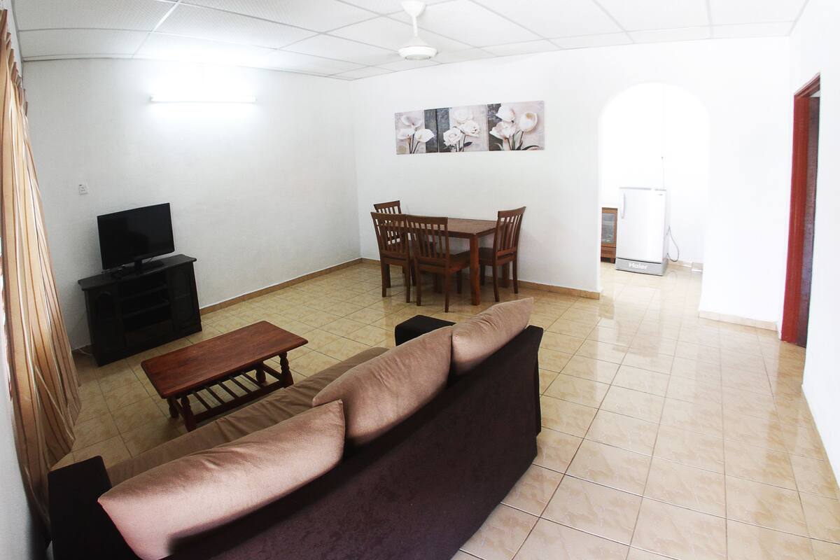 A living room with a sofa, a coffee table, a TV, a dining table with chairs, and a refrigerator.