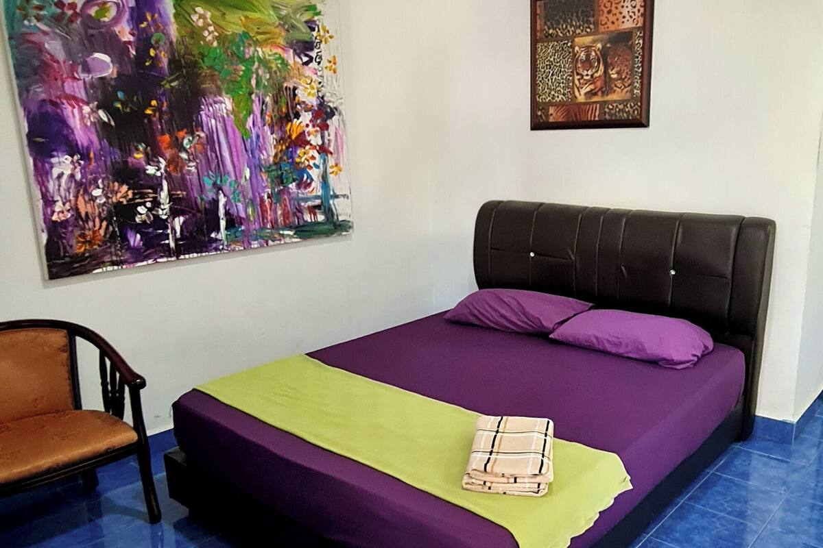 A bedroom with a bed, a chair, a colorful painting, and an air conditioning unit.
