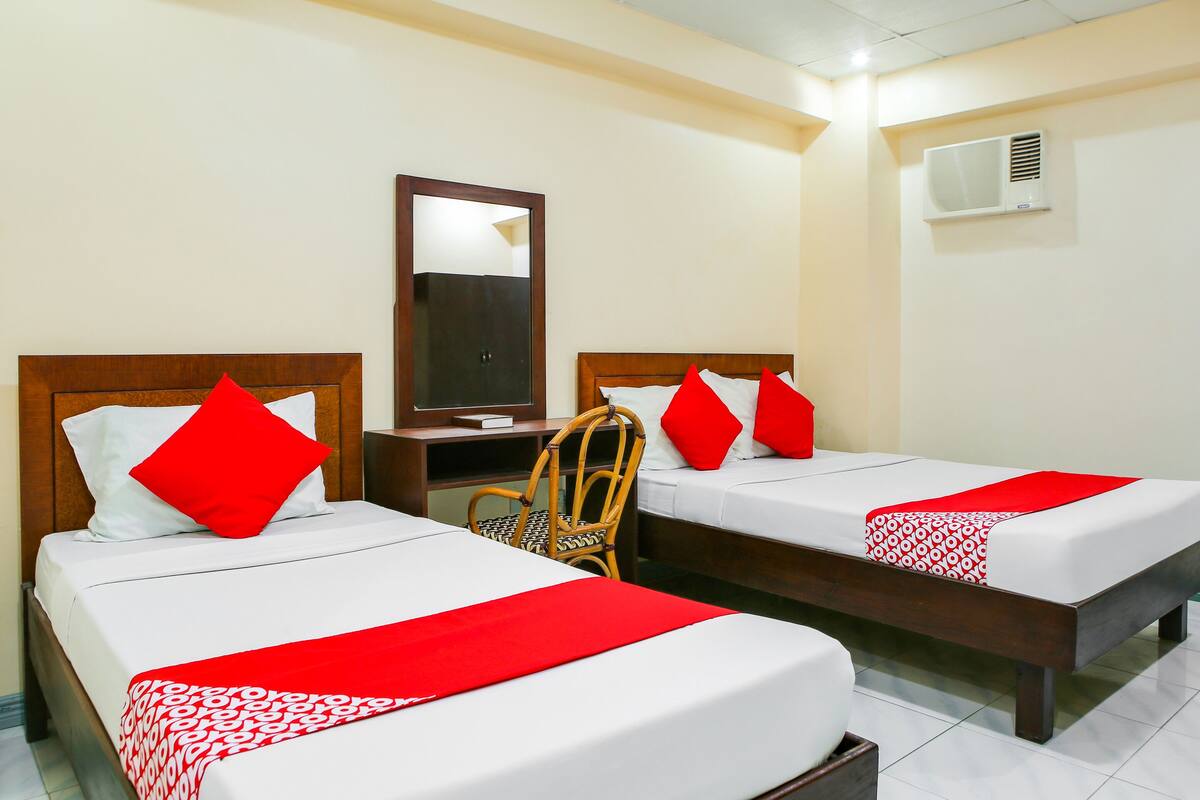 Habitación de hotel con dos camas individuales, cada una con almohada roja y colcha estampada. Se ve una silla de madera y un escritorio con espejo.