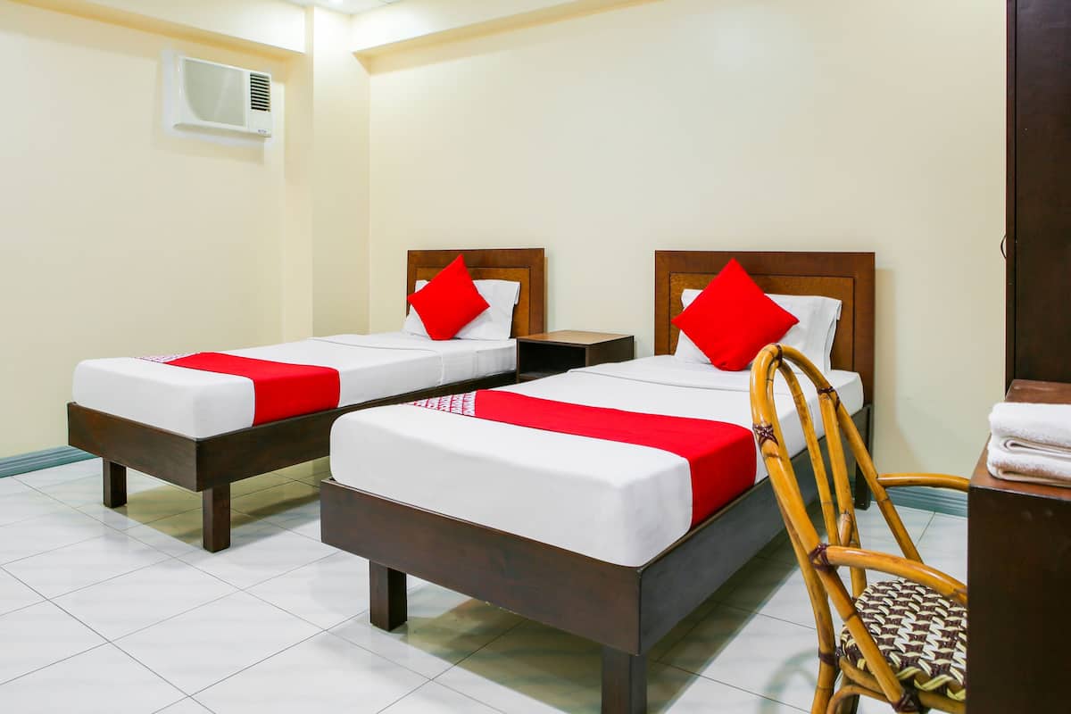 Habitación de hotel con dos camas individuales, cada una con almohada roja y ropa de cama blanca. Hay una silla de madera y una mesita con una toalla.