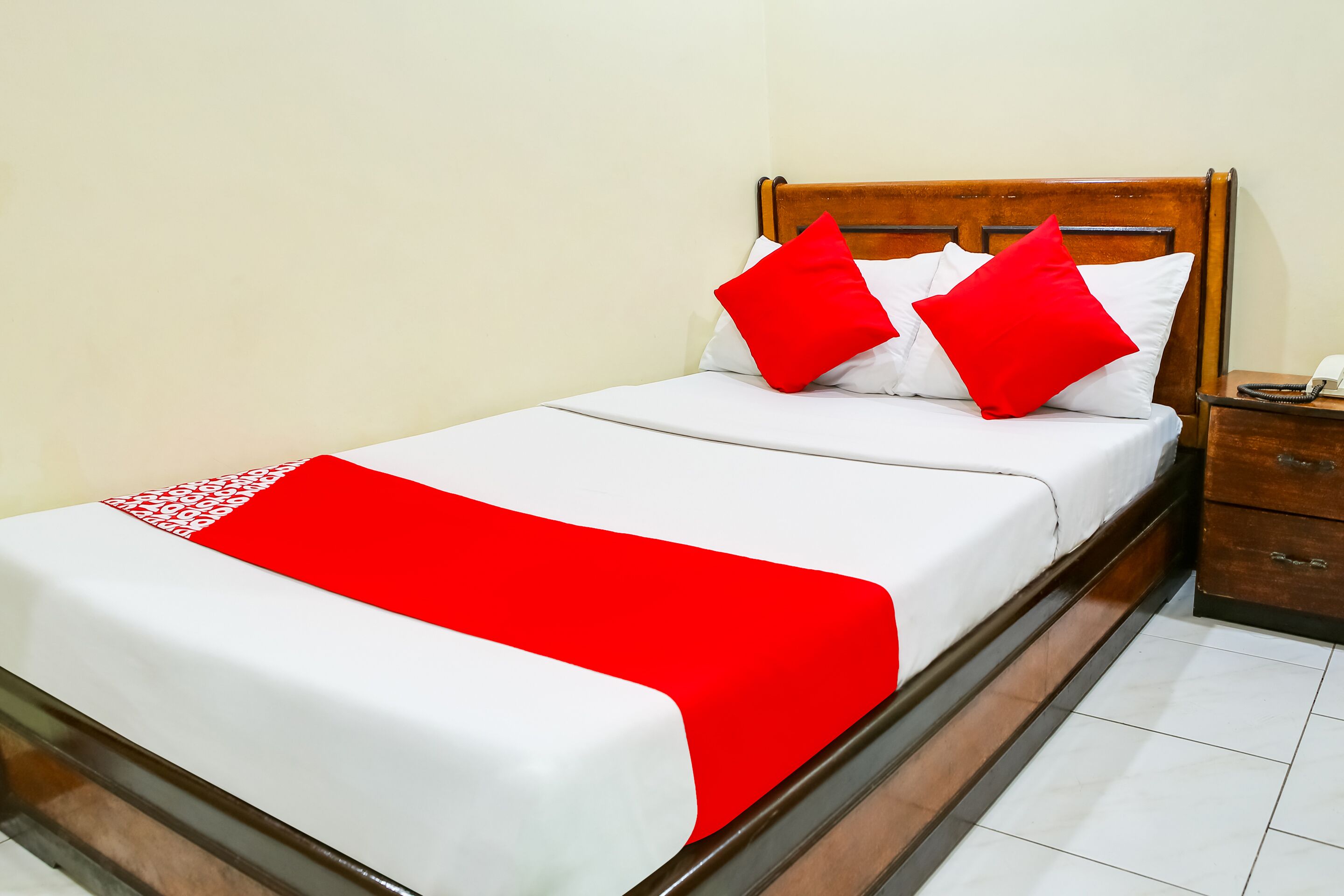 A neatly made bed with white and red bedding, a wooden headboard, and a bedside table with a telephone.