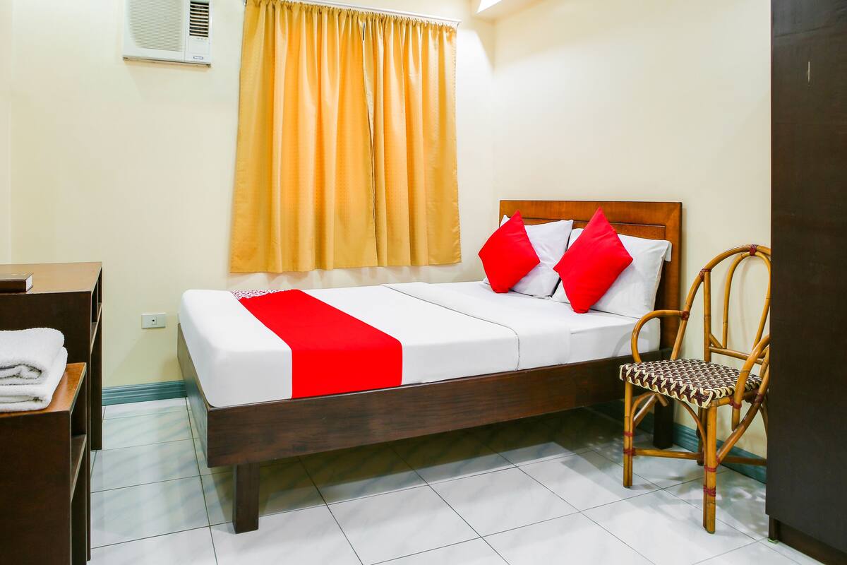 Una cama bien hecha con una colcha blanca y roja, un cabecero de madera y una almohada roja. Hay una silla de mimbre con asiento y respaldo trenzados, y una pequeña mesa de madera con una toalla blanca doblada.