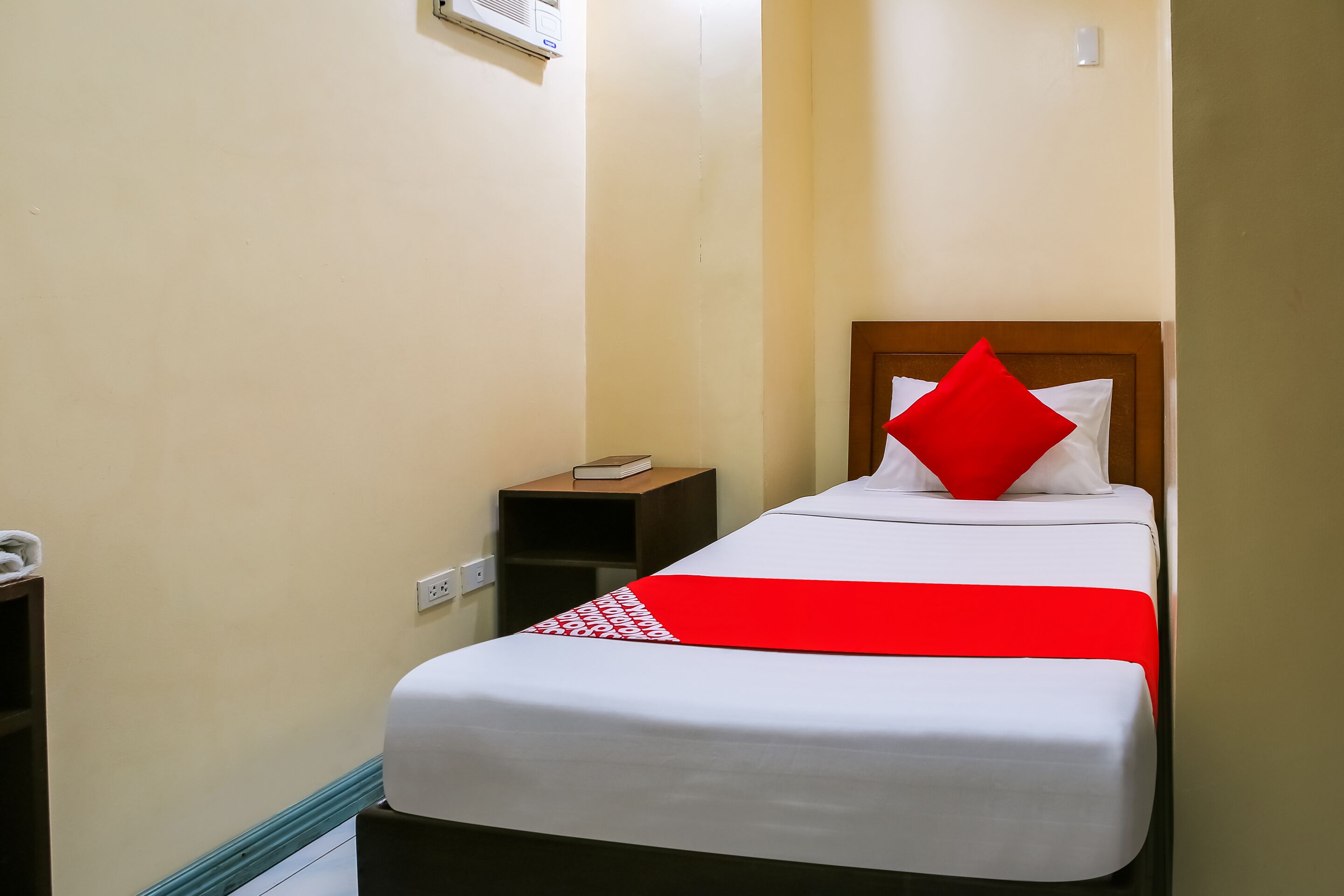 Habitació d'hotel estreta amb un llit individual, una tauleta de nit i una petita prestatgeria.