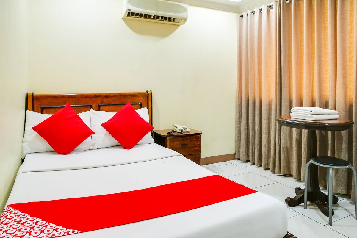 Una cama bien hecha con una colcha roja y blanca, un cabecero de madera y dos almohadas rojas. Se observa una mesita de noche con un teléfono y una toalla doblada. Junto a la cama hay un taburete pequeño, y una cortina está parcialmente abierta.