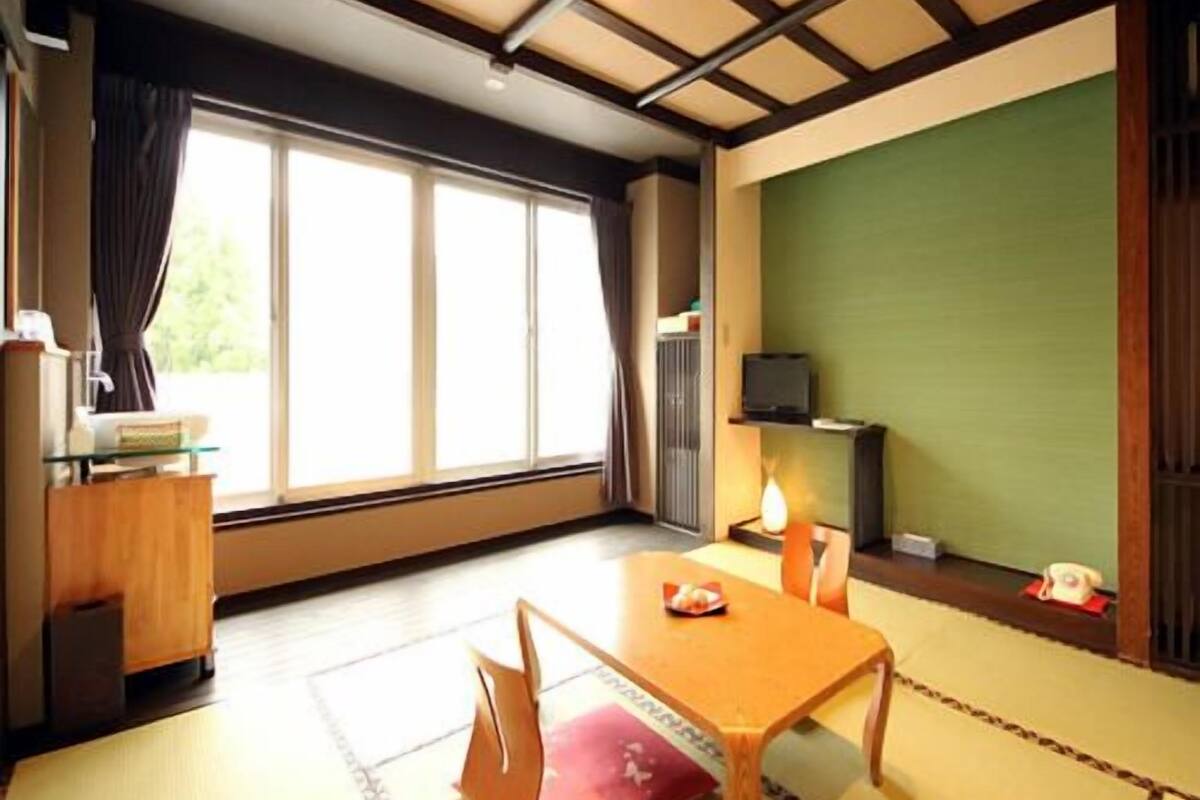 Ein traditioneller japanischer Raum mit Tatami-Boden, einem niedrigen Holztisch mit Kissen, einem Fernseher und einer Wand in Grüntönen.