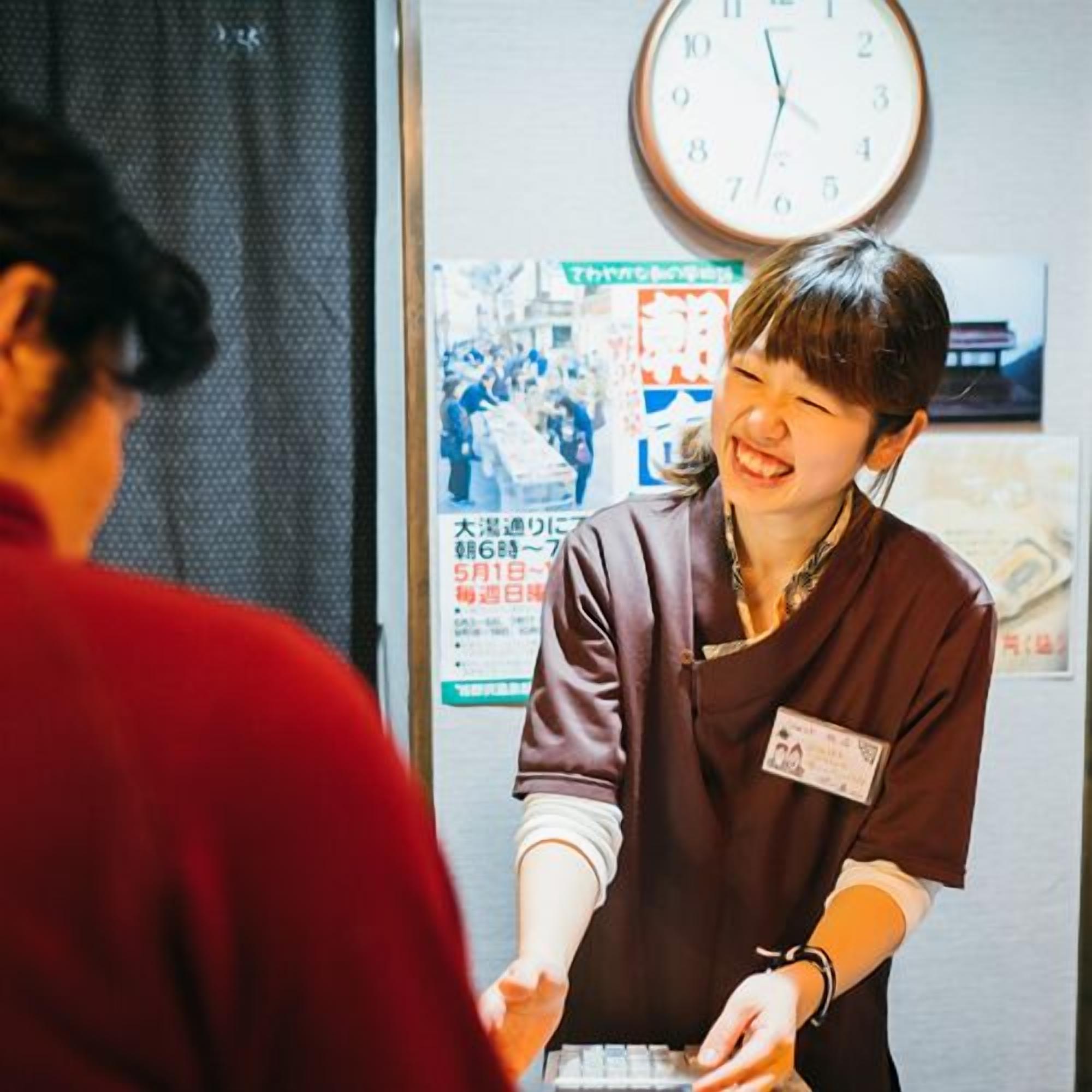 A person in a brown uniform handing over a card to another person. There is a clock on the wall and a poster with Japanese text.