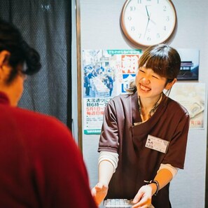 A person in a brown uniform handing over a card to another person. There is a clock on the wall and a poster with Japanese text. - Kawaichiya Ryokan (Nozawaonsen)