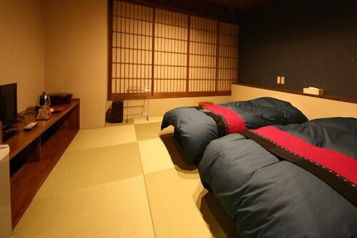 Ein traditioneller japanischer Raum mit Tatami-Böden, einem niedrigen Holztisch, einem Fernseher und einem Futonbett mit roter Akzentfarbe.