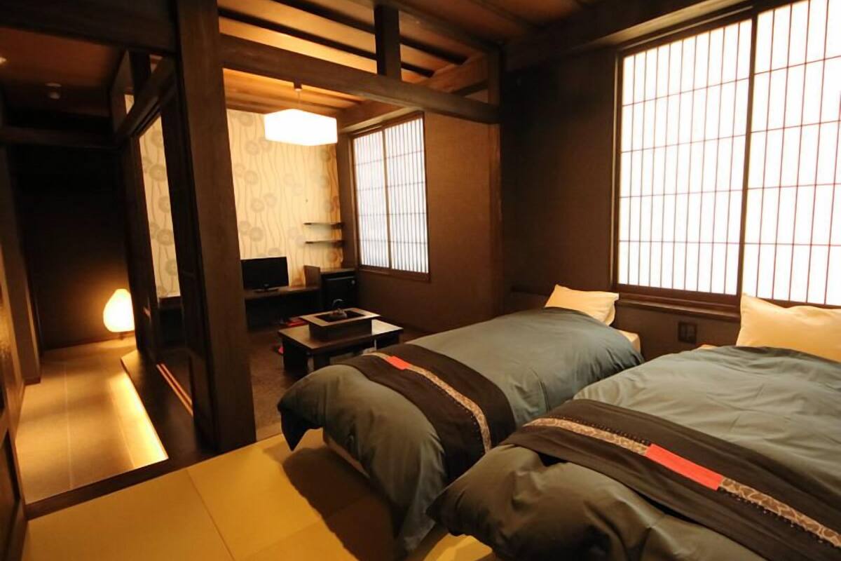 Ein traditioneller japanischer Raum mit Tatami-Bodenbelag, zwei Betten mit dunkler Bettwäsche, einem kleinen Tisch und einem Fenster mit vertikalen Jalousien.
