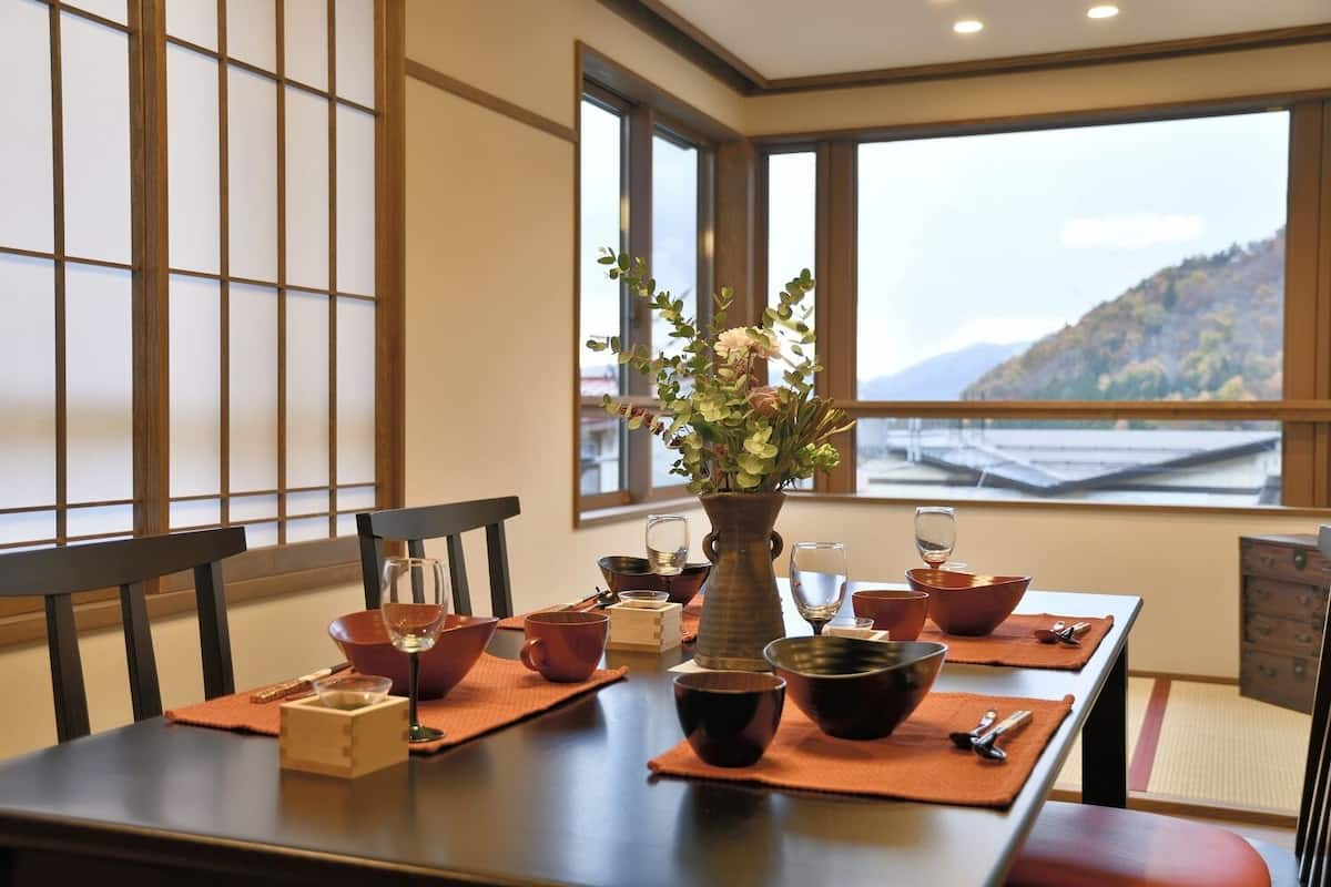 A dining table set with bowls, cups, and a vase of flowers, with a view of a mountain through large windows.