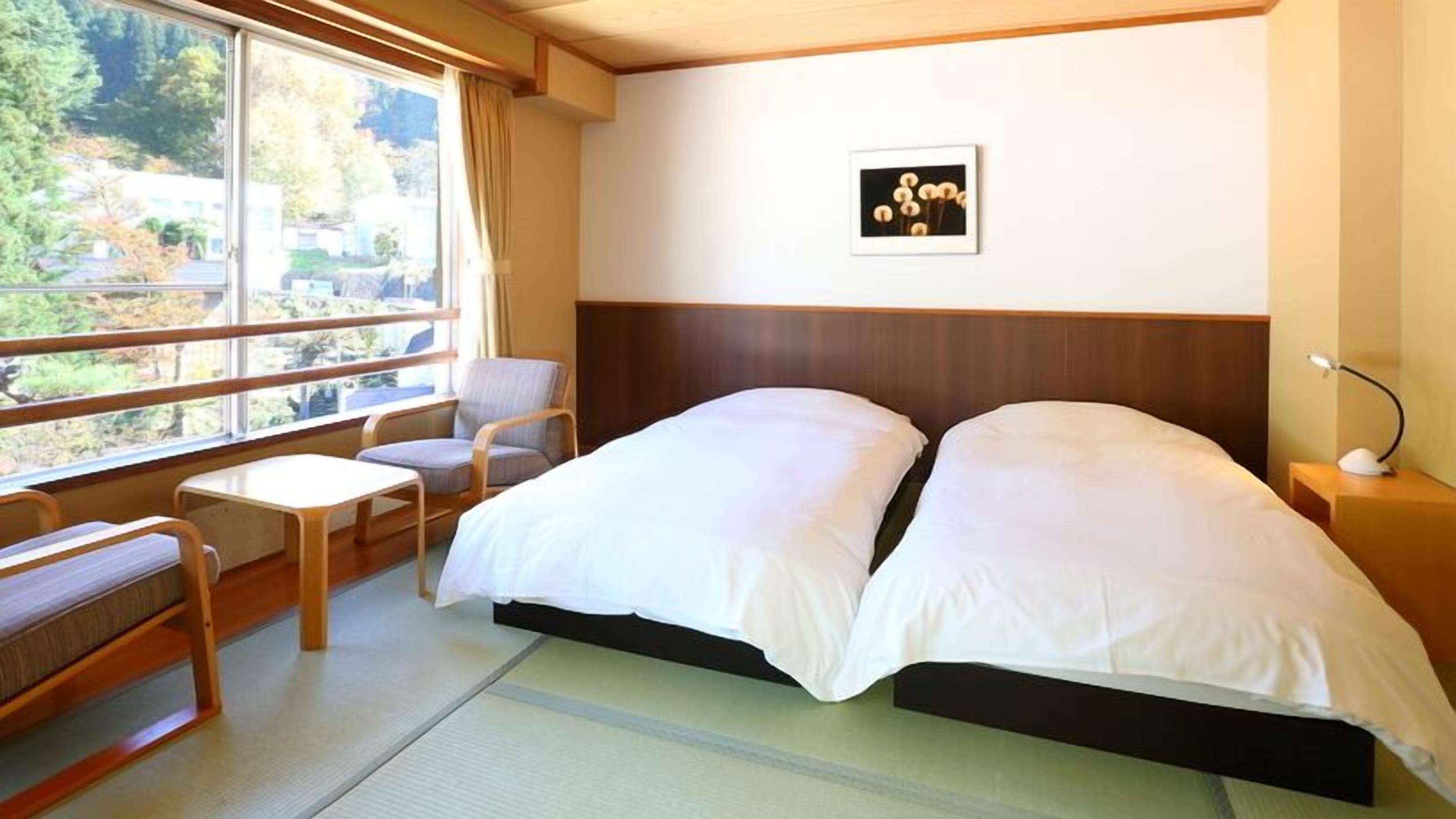 Habitación de hotel con cama, una silla, una mesita pequeña, una mesita de noche y un cuadro en la pared.