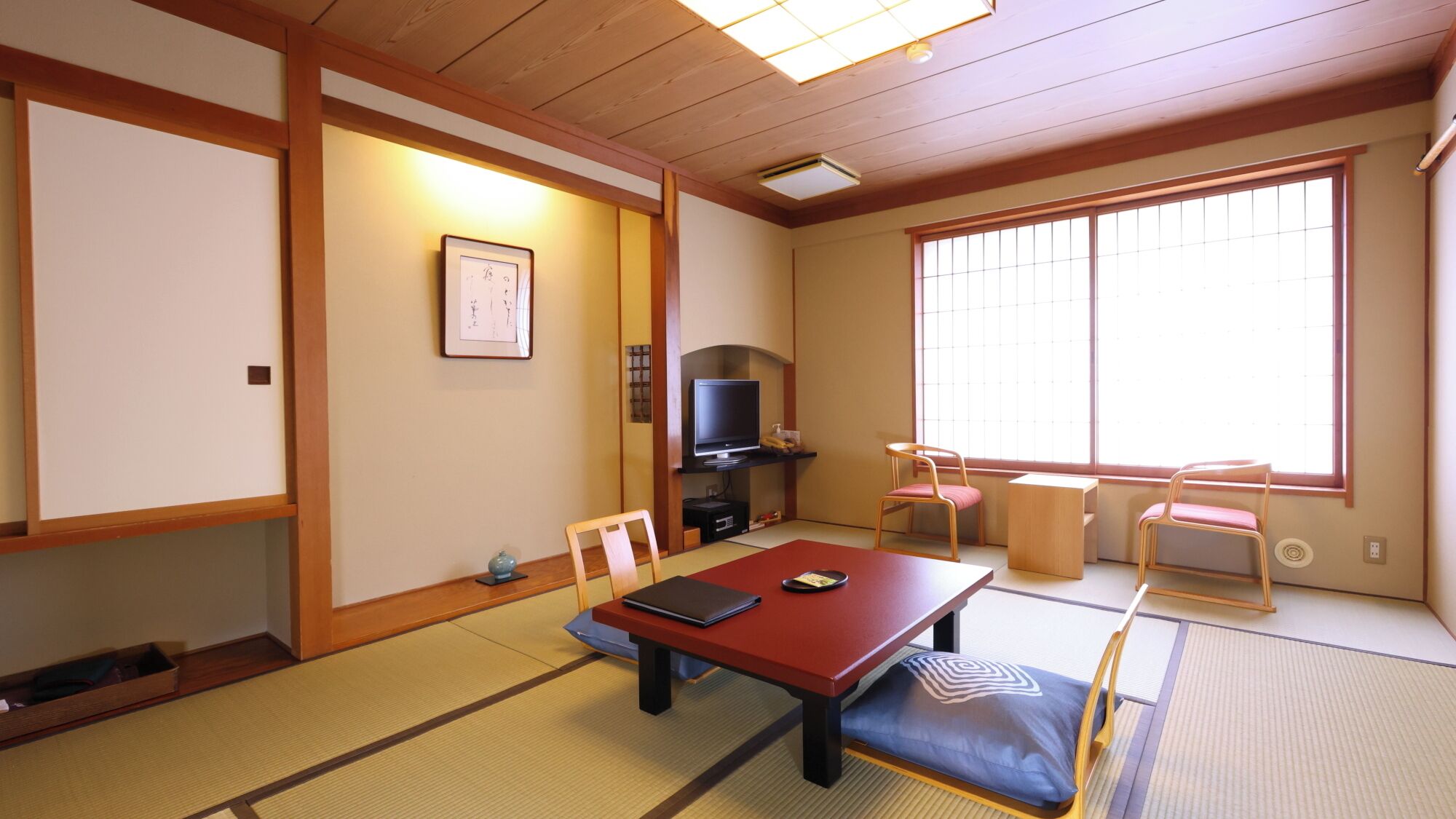 Una habitación japonesa tradicional con suelo de tatami, una mesa baja, sillas, un televisor y un ventanal grande.