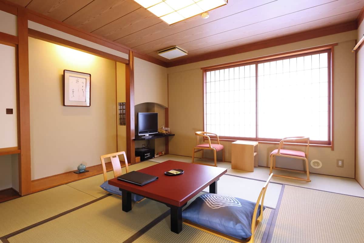 Una habitación japonesa tradicional con suelo de tatami, una mesa baja, sillas, un televisor y un ventanal grande.