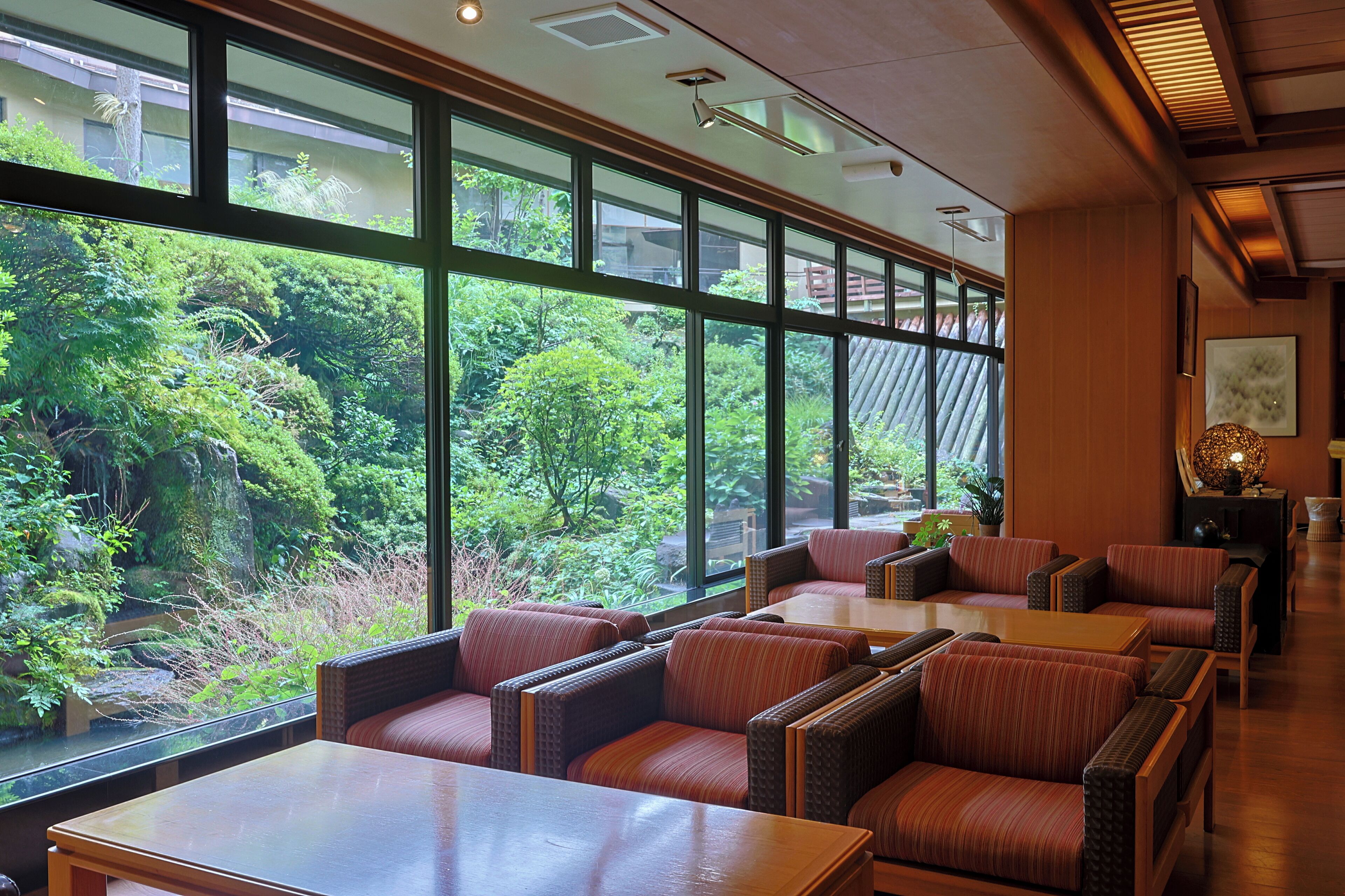 A restaurant with large windows offering a view of lush greenery. There are multiple seating areas with cushioned chairs and tables.