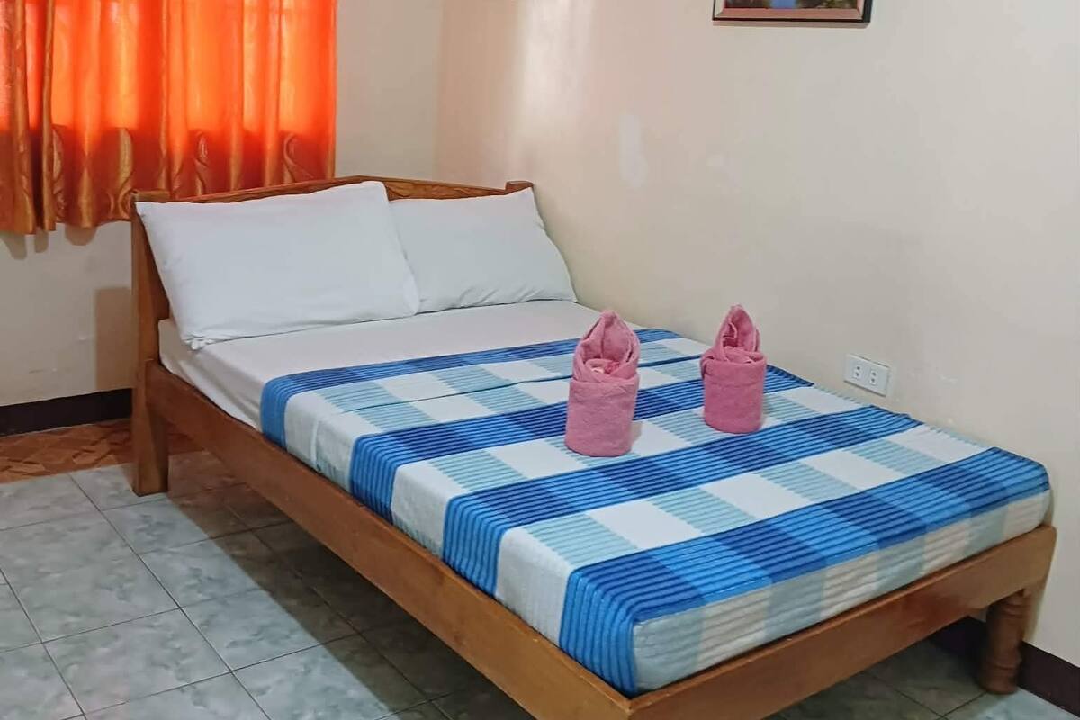 Una cama individual con colcha azul y blanca a cuadros, dos toallas rosas y un cuadro con un paisaje en la pared.