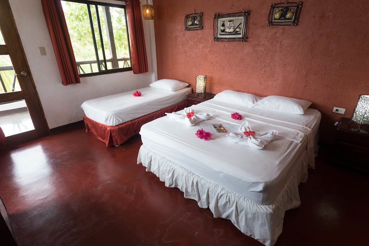 A hotel room with two beds, a wooden floor, a window with curtains, and wall-mounted pictures.