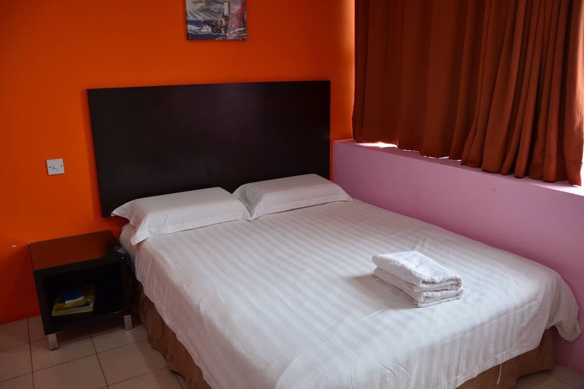 Un lit simple avec du linge de lit blanc et une serviette pliée, une tête de lit noire et une table de chevet avec un livre.