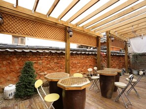 An outdoor patio with wooden tables and chairs, a stone wall, and a wooden pergola. - TREGA Museum Guesthouse (Jeonju)