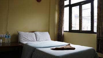 Una cama bien tendida con sábanas blancas y una manta marrón, una mesita de noche con botellas de agua y una ventana con cortinas.