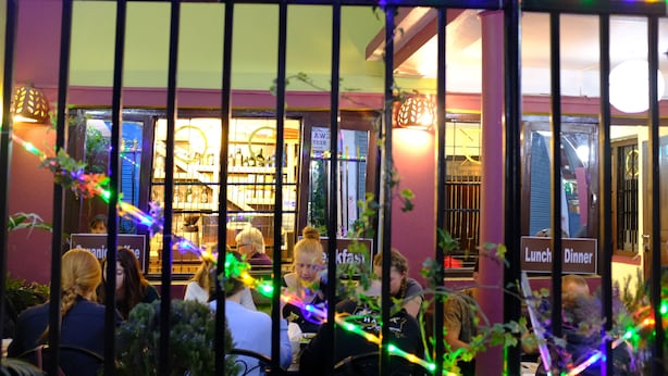 Ein Restaurant mit Außenbereich, einem Schild, das 'Mittagessen' und 'Abendessen' anzeigt, und dekorativer Beleuchtung.