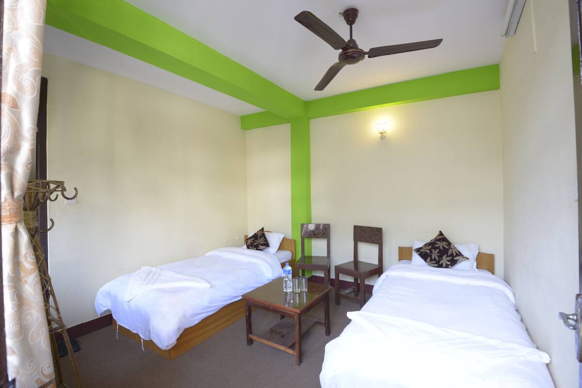A hotel room with two single beds, a small table with water bottles, a ceiling fan, and a green accent wall.