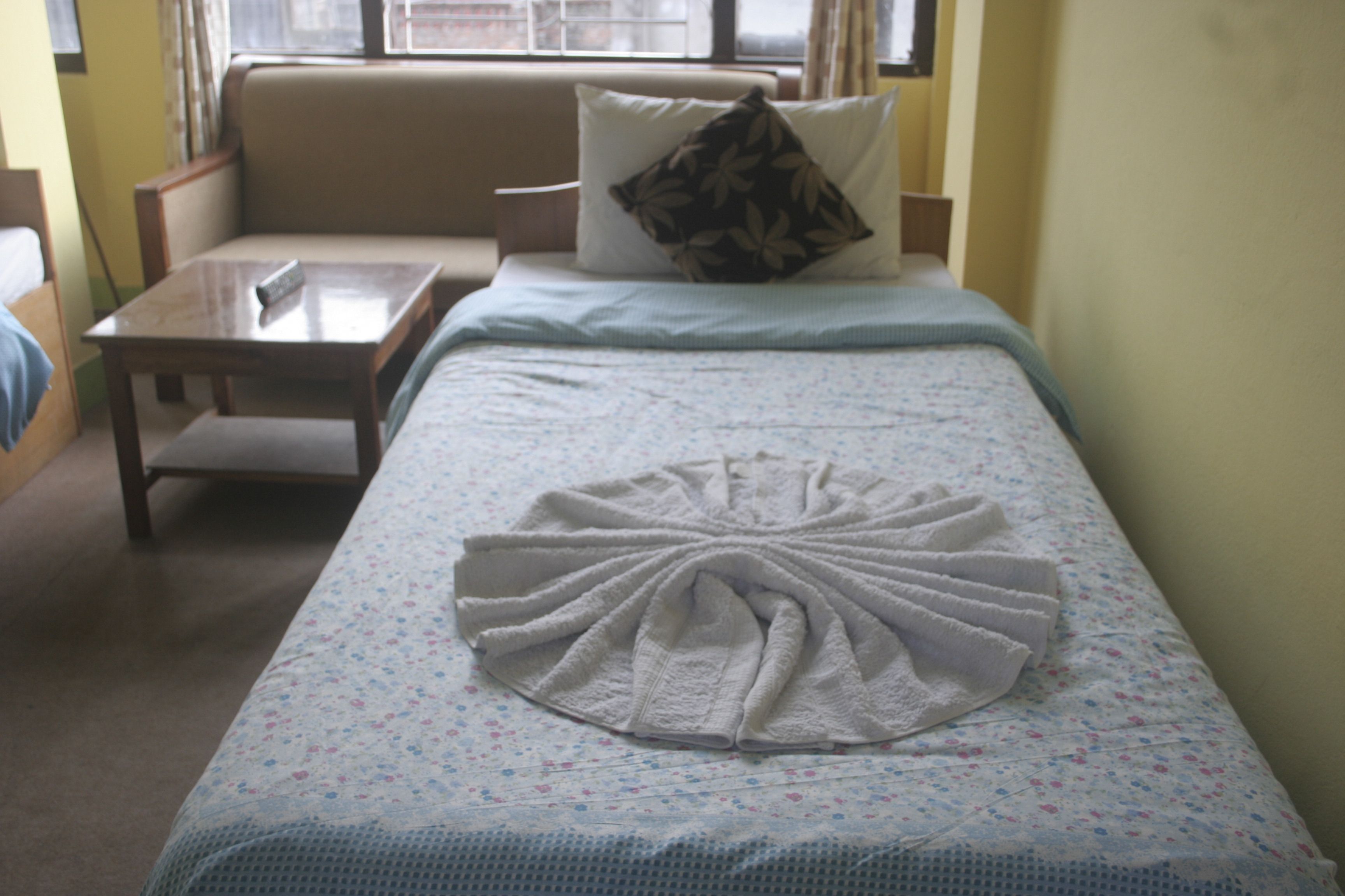 A neatly made bed with a decorative towel, a small bedside table, and a sofa in a room with yellow walls.