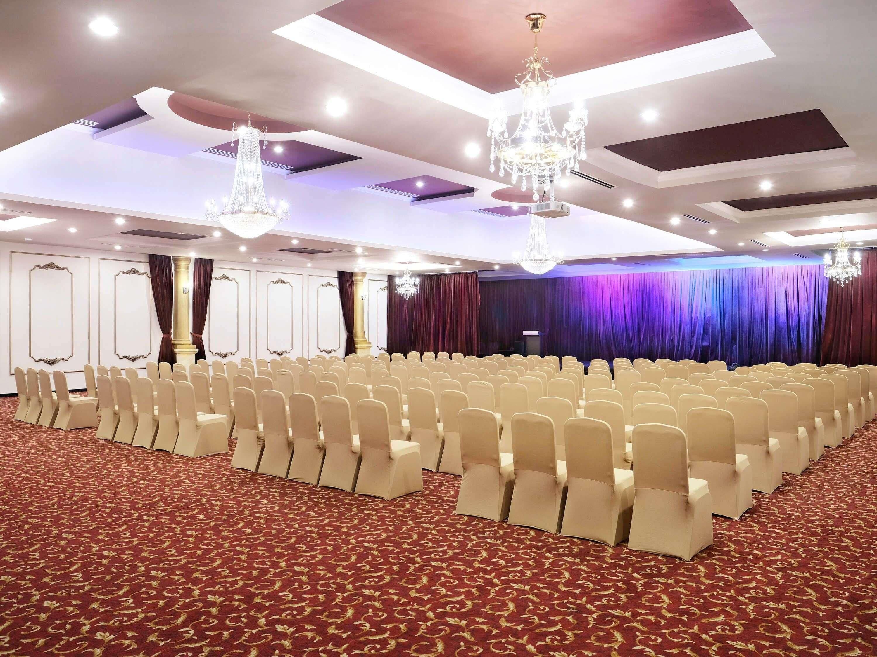 Un salón de conferencias con filas de sillas, un escenario con cortinas, candelabros y una alfombra estampada.