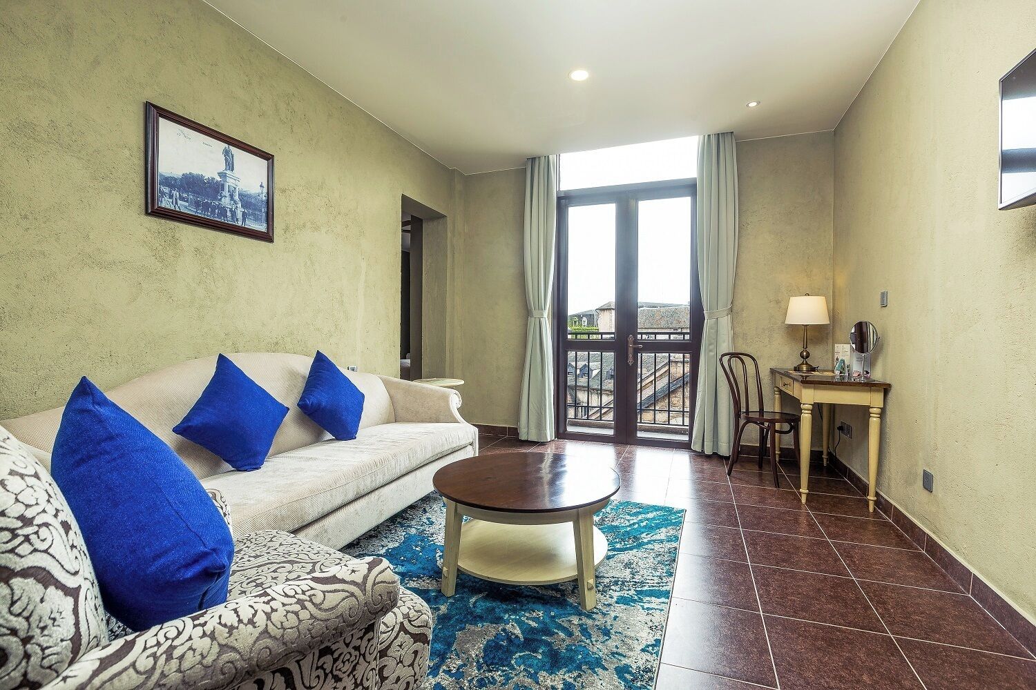 A living room with a sofa, armchair, and coffee table. There is a balcony with a view of buildings and a door leading to another room.