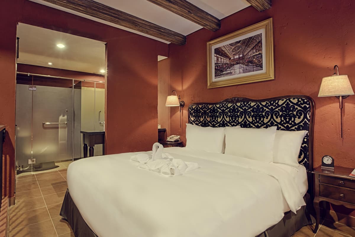 Een hotelkamer met een groot bed, een nachtkastje, een lamp, een inlijsting foto aan de muur, en een badkamer met een glazen deur.
