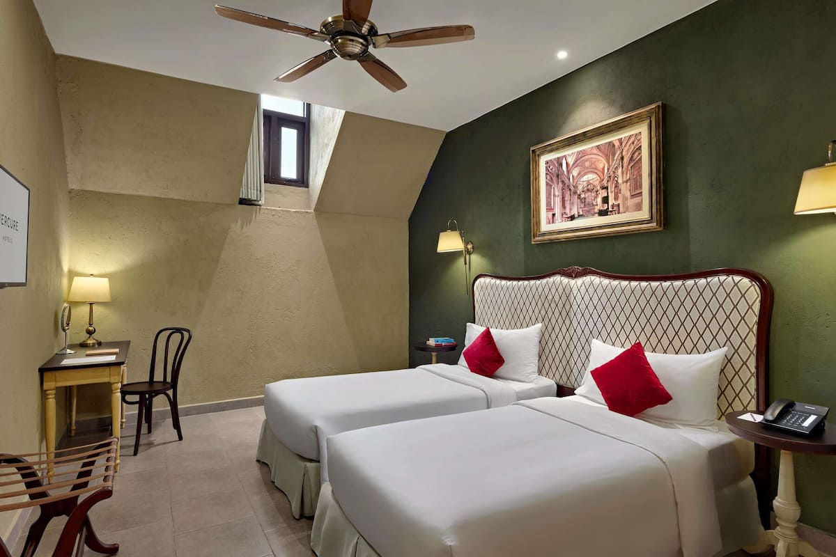 Een hotelkamer met twee bedden, een bureau met lamp, een stoel, een plafondventilator en een schilderij aan de muur.