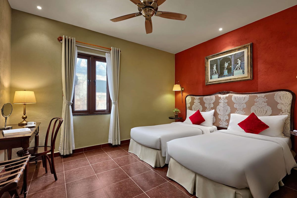 Een hotelkamer met twee bedden, een plafondventilator, een raam met gordijnen, een bureau met lamp, een stoel en een lijst met een afbeelding aan de muur.