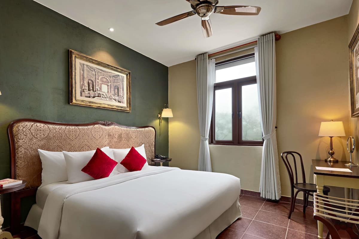 Hotelkamer met een groot bed, een plafondventilator, een raam met gordijnen, een bureau met lamp en een lijst met een afbeelding aan de muur.