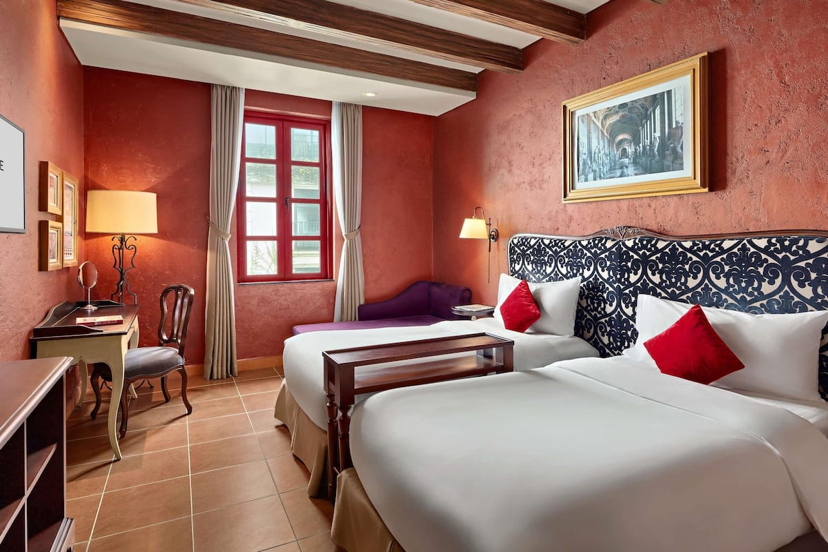 Een hotelkamer met twee bedden, een bureau, een stoel, een raam met gordijnen en een foto in een lijst aan de muur.