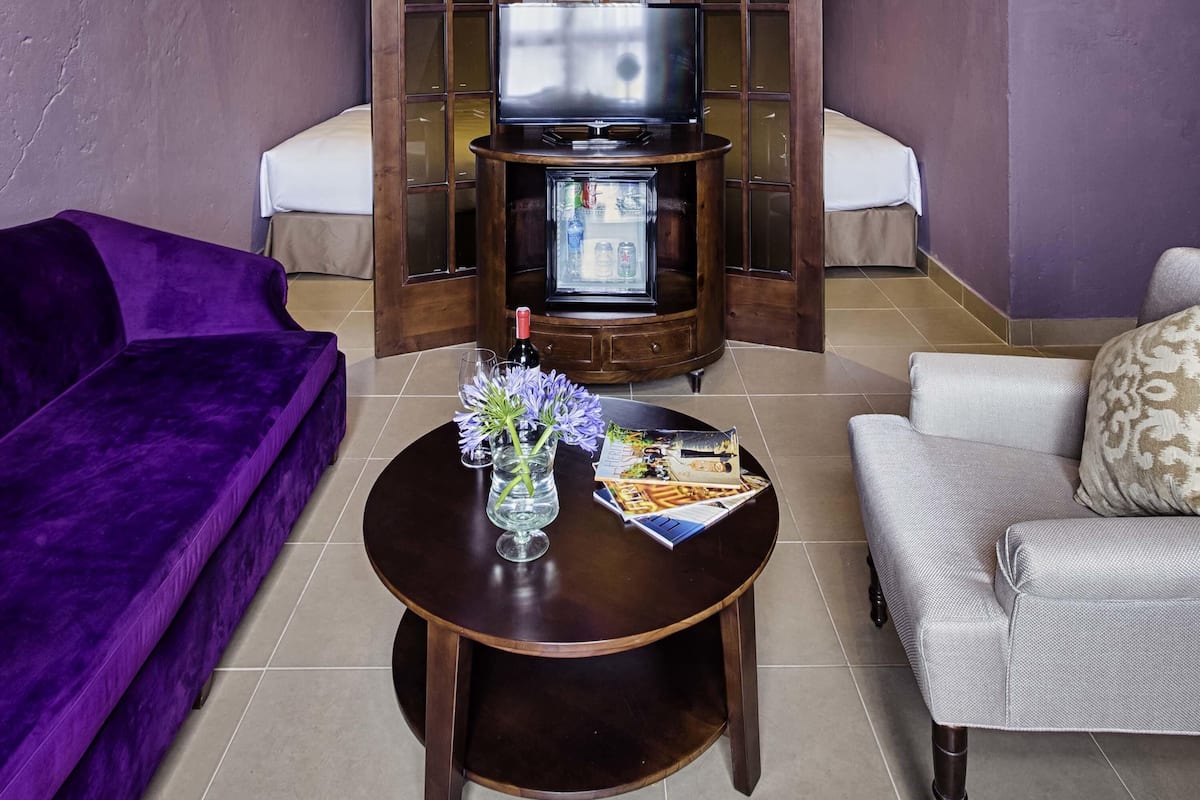 Un salon avec un canapé violet, un canapé gris, une table basse ronde en bois sur laquelle reposent un magazine et une bouteille de vin, et un téléviseur posé sur un meuble en bois.