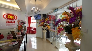 Zona de recepción de hotel con mostrador, candelabro, asientos rojos y un amplio ventanal con flores.