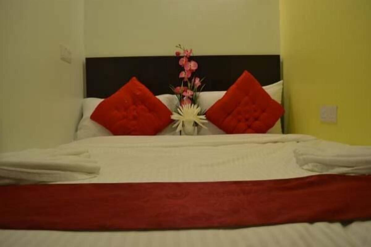 Una cama bien tendida con almohadas rojas y un edredón blanco, un pequeño florero blanco con flores y un cabecero oscuro.