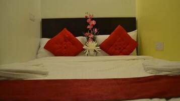 Una cama bien tendida con almohadas rojas y un edredón blanco, un pequeño florero blanco con flores y un cabecero oscuro.