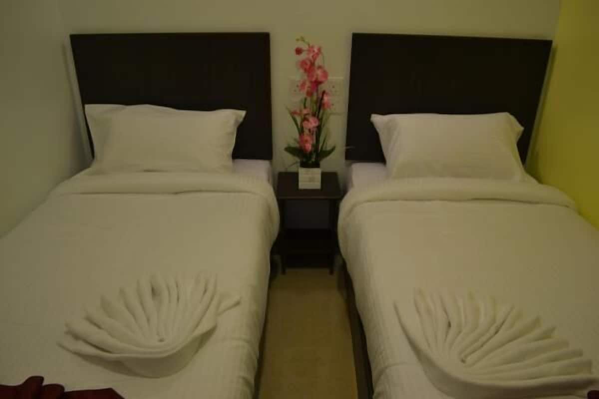 Habitación de hotel con dos camas, cada una con cubrecama y almohadas blancas. Entre las camas hay una mesita de noche con un jarrón de flores rosas.