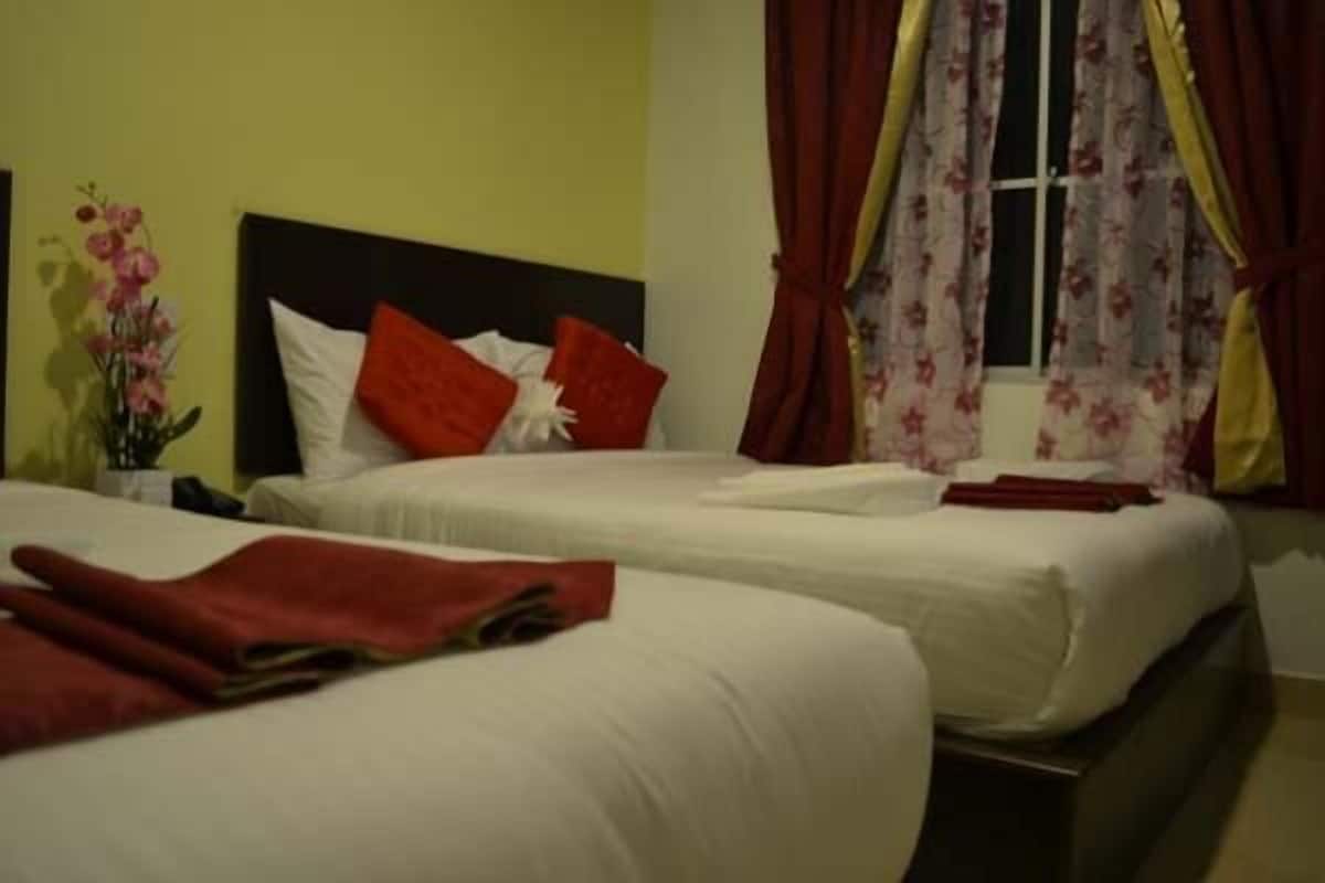 Habitación de hotel con dos camas, cada una con ropa blanca y almohadas rojas. Se ve una ventana con cortinas florales, y un jarrón con flores a la izquierda.