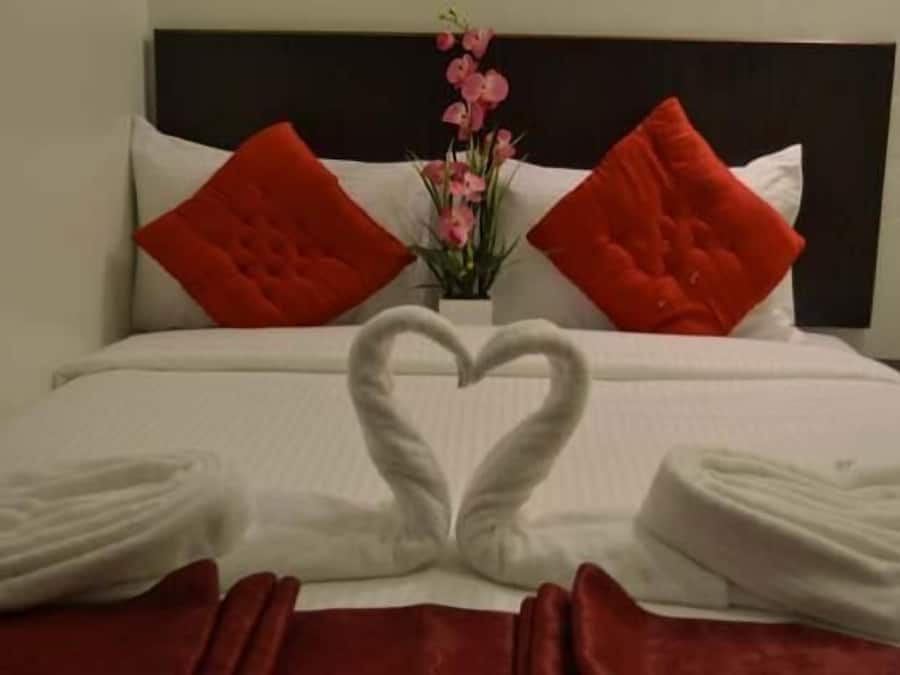 A neatly made bed with a heart-shaped towel arrangement, red pillows, and a vase of pink flowers.