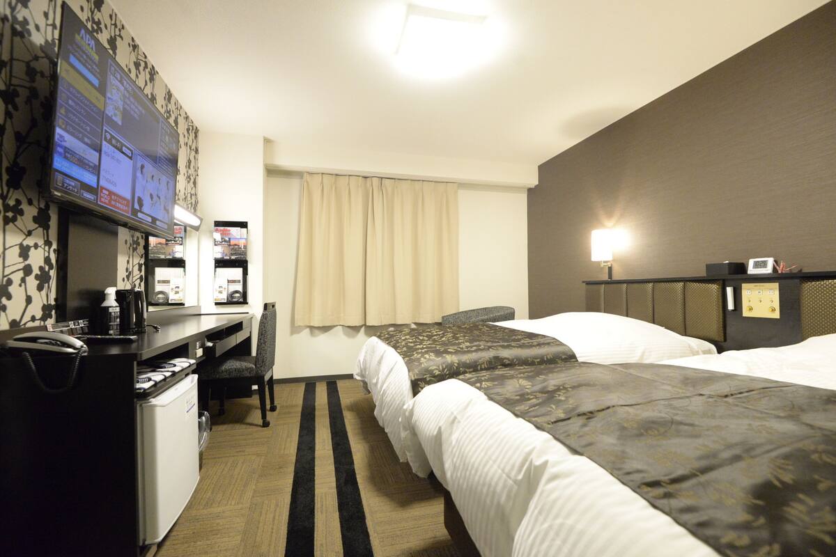 Habitación de hotel con dos camas, un escritorio con computadora, una silla y un televisor mostrando información del hotel.