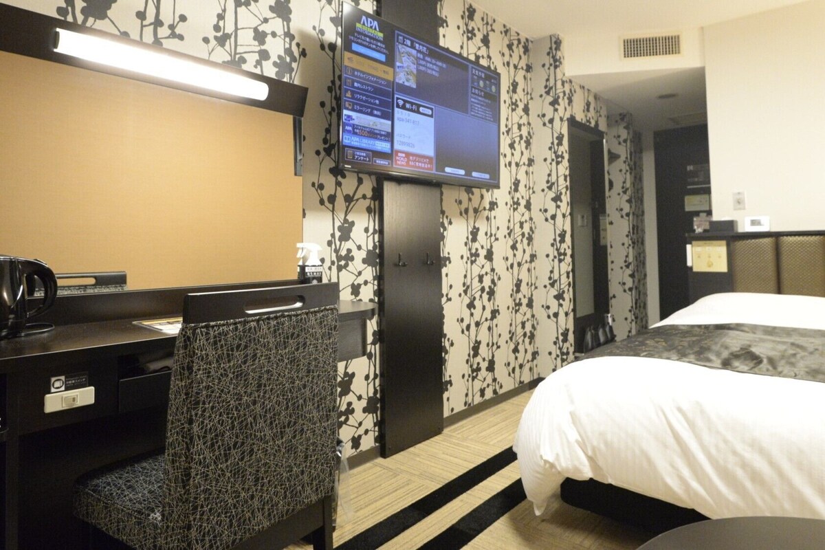 Une chambre d’hôtel comprenant un lit, un bureau avec une chaise, une télévision et un mur orné d’un motif floral.