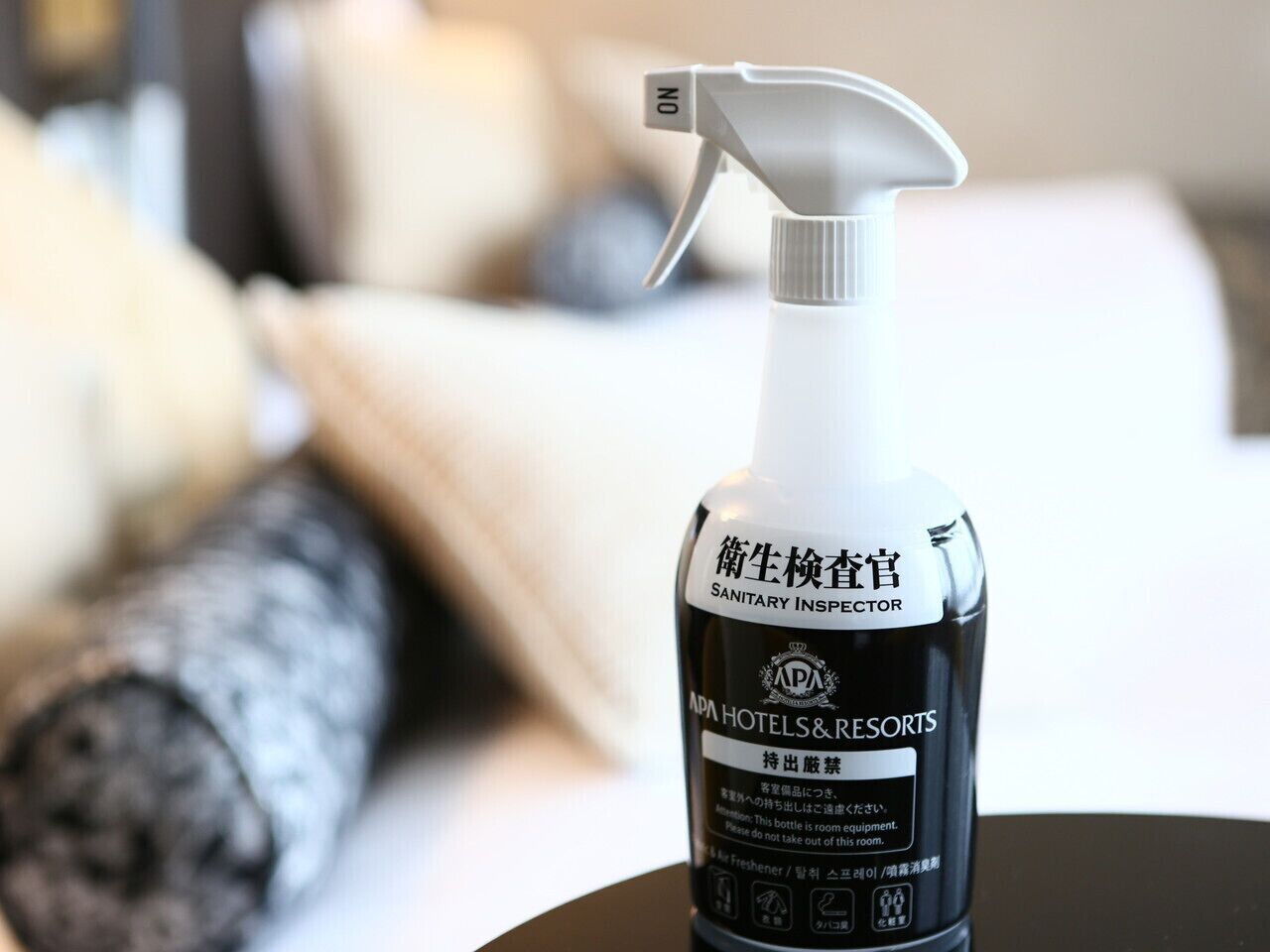 A bottle of hand sanitizer on a table with a blurred background of a bed and pillows.
